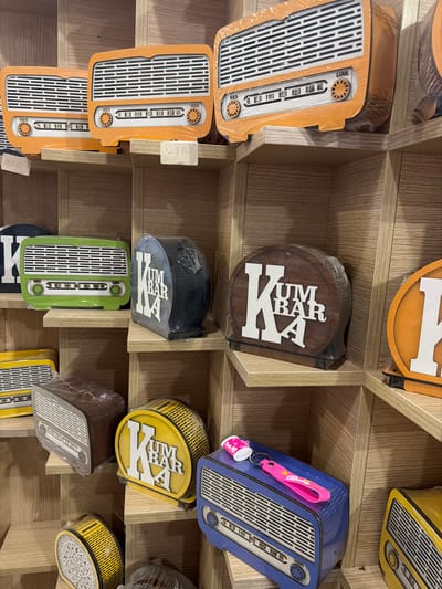 This image displays a close-up, eye-level view of a retail display shelf filled with decorative items shaped like vintage radios. The items are arranged on wooden shelves in a store. In the foreground, on the bottom shelf, are two circular wooden signs with the letters "KUM BAR A" on them, one yellow and one brown. To the left of these, on the same shelf, is a brown rectangular item that also resembles a vintage radio, wrapped in plastic. On the shelf above, a green, a black, and an orange vintage radio-shaped item are visible. The orange items on the top shelf have price tags. The overall scene suggests a retail environment showcasing novelty or decorative items. The location is in Acıpayam, Turkey.