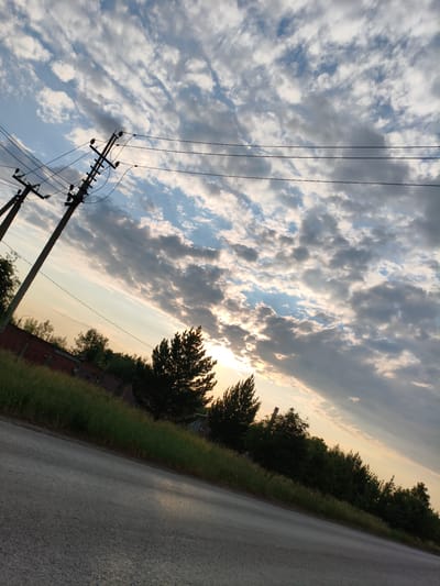 This media file captures an outdoor scene with a dramatic sky and elements of a rural or semi-rural setting. The primary subjects include power lines supported by utility poles, a road in the foreground, and a line of trees and vegetation bordering the road. The sky is filled with a complex cloud formation, with patches of blue sky visible between the clouds. Sunlight is breaking through the clouds in the upper right portion of the image, creating rays of light that extend downwards.

The time of day appears to be late afternoon or early evening, given the warm, soft lighting and the angle of the sun. The weather seems to be partly cloudy, with the clouds creating dynamic patterns and shadows across the sky. The location cues, such as the style of the power poles and the vegetation, suggest a setting in Металлплощадка, Russia. The overall mood of the image is somewhat serene and contemplative, enhanced by the natural beauty of the sky and the quiet landscape.

There are no people visible in the image, and no specific activity or event is taking place. The focus is on the landscape and the atmospheric conditions. Notable details include the texture of the road surface, the green grass and trees, and the details of the insulators on the power poles. The image is angled, with the road and horizon running diagonally from the bottom left to the upper right, which adds a sense of depth and perspective. There is no visible text in the image.