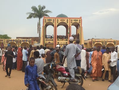 In Kafanchan A, Nigeria, a large group of people, mostly men, are gathered outside a building with a green dome and three arches, possibly a palace. A palm tree stands to the left of the building. People are on the building's upper level. Many of the people are wearing traditional African clothing and head coverings. A man stands on a red motorcycle. Another motorcycle is parked nearby, and a young boy in a blue robe is looking at the camera in the foreground. The weather appears overcast.