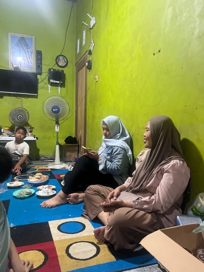 The image depicts an indoor gathering likely in a residential setting in Wedarijaksa, Indonesia. Two women, both wearing hijabs, are seated on a colorful rug on the floor. The woman on the right is wearing a pink top and brown hijab, and is smiling while looking to her right. The woman on the left is wearing a denim jacket over a white shirt and a light blue hijab, and is looking down at her phone. In the foreground, plates of food and drinks are laid out on the rug. To the left, a young boy in a white t-shirt is sitting on the floor, looking to his right. In the background, a wall painted a bright lime green dominates the scene. Various objects are visible on the wall, including a framed picture, speakers, a clock, and a ceiling fan. Two standing fans are also present in the room. The lighting appears to be artificial, creating shadows on the wall. The overall atmosphere suggests a casual social gathering.