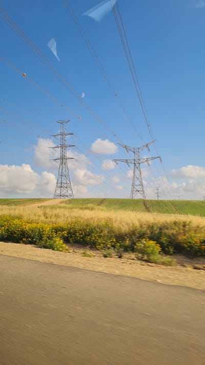 The image shows a view of power lines and a field of yellow flowers next to a road, likely taken from a moving vehicle. The sky is blue with scattered clouds. The power lines run parallel to the road and field. There are orange markers on the lines. The location is likely within or near the Bnei Shimon Regional Council in Israel, given the presence of power lines and agricultural fields which are typical of this area.