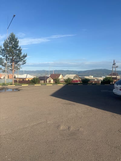 The image depicts an outdoor scene in Gusinoozyorsk, Russia. In the foreground, there is a large expanse of asphalt, with a shadow cast across it from an unseen object to the right. To the left of the asphalt, there are puddles of water collected on the ground, bordered by a curb with yellow and black markings. A tall pine tree stands to the left, with a lamppost nearby. In the middle ground, a row of houses with various roof types and fences are visible. Beyond the houses, a hazy landscape of hills and mountains stretches to the horizon. Two industrial chimneys with smoke rising from them are also visible in the distance. A white car is parked on the right side of the frame, partially visible. The sky is clear and blue with a few wispy clouds.