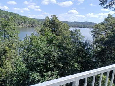The image shows a view across a body of water, likely a lake, surrounded by dense green forests. In the foreground, there is a light-colored metal railing, indicating a viewpoint or balcony. The water is calm and reflects the blue sky dotted with white clouds. The trees on the hillsides are a mix of coniferous and deciduous varieties, creating a lush, natural landscape. The scene appears to be a serene outdoor setting. The location is the Mácha Lake in Doksy, Czechia.