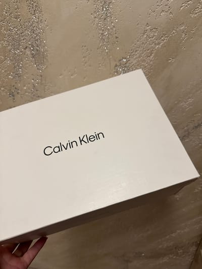 A close-up, slightly angled shot shows a white Calvin Klein shoebox with the logo clearly visible in black text. The box is resting on a textured, taupe-colored surface, possibly a wall or a textured tabletop. Part of a hand is visible at the bottom left corner, holding the box. The background has a rough, uneven texture with some reflective, glitter-like elements. The lighting appears to be indoor ambient light. There is no discernible location in Rogosh, Bulgaria.