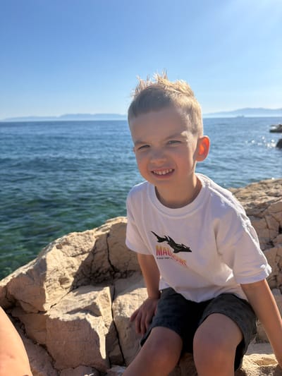 An image, originally oriented horizontally, captures a joyful young boy posing by the sea in Kostrena, Croatia, under a bright, clear sky. The boy, with light brown hair and a wide, beaming smile, looks directly at the camera. He appears to be in his early elementary school years. He is dressed in a white short-sleeved t-shirt featuring a black silhouette of a shark or similar marine creature, with the text "MAUI AND SONS" and "SINCE 1980" visible below it. He also wears dark shorts.
He is seated on large, light-colored rocks, illuminated by strong sunlight, which creates distinct shadows and highlights their rugged texture. Behind the boy and stretching into the background is a vast expanse of calm, blue water, likely the Adriatic Sea, with distant land or mountains faintly visible on the horizon. The bright light and clear conditions indicate a sunny day, possibly morning or late afternoon. The boy's expression conveys happiness and enjoyment in his surroundings.