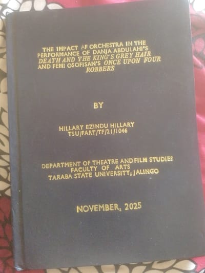 The image shows the cover of a dark blue book with gold lettering. The title appears to be "THE IMPACT OF ORCHESTRA IN THE PERFORMANCE OF DANJIA ABDULAHI'S DEATH AND THE KING'S GREY HAIR AND FEMI OSOFISAN'S ONCE UPON FOUR ROBBERS". Below the title, it states "BY HILLARY EZINDU HILLARY TSU/FART/TF/21/1046". Further down, it reads "DEPARTMENT OF THEATRE AND FILM STUDIES FACULTY OF ARTS TARABA STATE UNIVERSITY, JALINGO". The date "NOVEMBER, 2025" is at the bottom. The book is lying on a surface with a patterned fabric, possibly a bedspread, with shades of pink, white, and brown. The image is taken from a slightly overhead angle. There is no indication of the city of Magajin Gari, Nigeria.