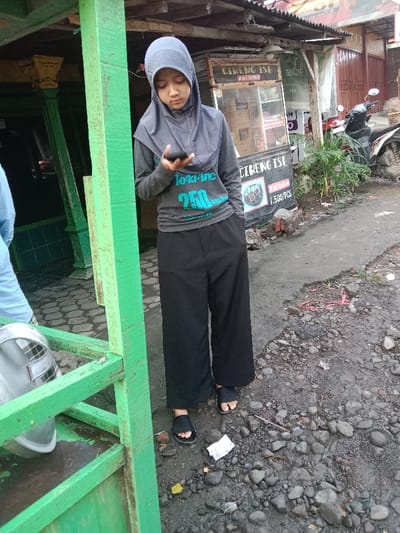 A woman wearing a hijab, a grey long-sleeved shirt with a turquoise logo, and wide black pants stands on a rocky, paved surface. She is looking at a dark smartphone in her hand. A green painted structure is to her left. In the background, there is a street food vendor with a sign reading "CIRENG ISI" and a parked motorcycle. The overall lighting suggests it is daytime.
