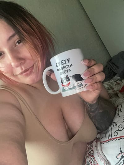 A woman is lying in bed, holding a white mug with a drawing of two cats on it. The mug also has text in Russian that reads "СУЕТУ НАВЕСТИ ОХОТА". She is wearing a beige bra. The bed is covered with a patterned duvet. The background appears to be a wall. The image was taken indoors. There is no information to determine the location in the city of Коробицыно, Russia.
