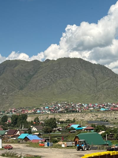 The image depicts a rural village nestled at the base of a large, green, and rocky mountain under a bright blue sky with scattered white clouds. The village consists of numerous colorful houses with red, blue, and green roofs. In the foreground, a dirt road is visible, with a red car parked on the left side. A blue tractor is positioned in front of a large green-roofed building. The environment appears to be a remote settlement in a mountainous region, possibly in or around the city of Onguday, Russia, given the architectural style of the houses and the mountainous landscape.