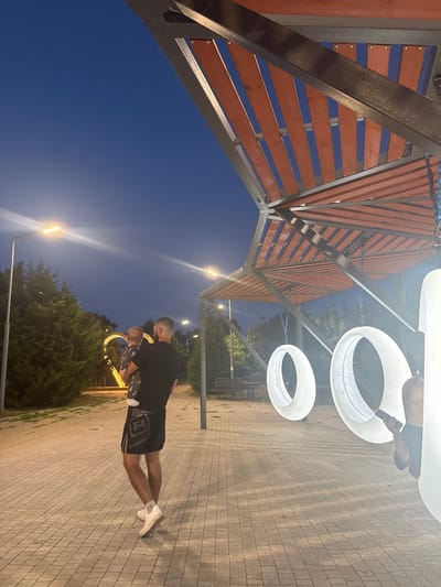 In Egorlykskaya, Russia, at dusk, a man holding a baby stands on a paved walkway. The man is wearing black shorts with white lettering and white sneakers. He is facing away from the camera, looking towards a large, illuminated heart-shaped structure in the distance. Behind him, a modern canopy with wooden slats is visible, with large, white, ring-shaped objects hanging from it. The sky is a deep blue, and streetlights illuminate the path. Green trees line the walkway.