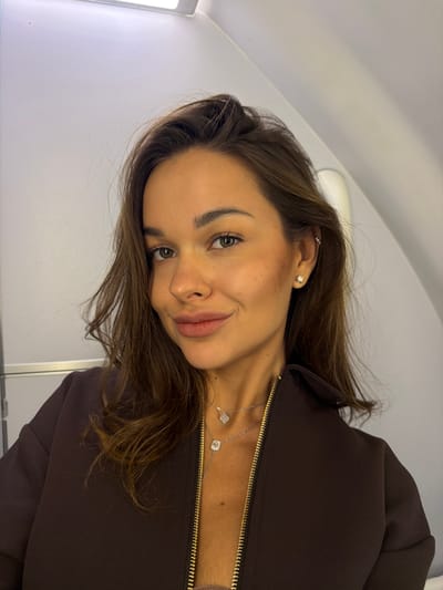 A young woman with brown hair is taking a selfie inside an airplane cabin. She is wearing a dark brown zippered jacket, a nude-colored top, and two silver necklaces. Her left ear has two piercings, with diamond stud earrings in both her upper and lower earlobe. She is smiling faintly, looking directly at the camera. The background shows the interior of an airplane, with white walls and a ceiling light. The location is within the city of Перепечино, Russia.