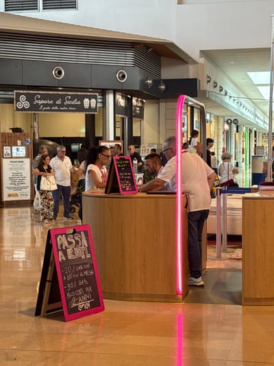 This vertically oriented, eye-level, full shot captures a busy indoor scene, possibly a food court or shopping mall, with people queuing and interacting with food vendors. The lighting is bright, suggesting daytime, and there are no visible signs of weather.
In the foreground, a man wearing a white t-shirt and dark jeans is leaning over a wooden counter, his face obscured. To his right, a woman in a white top and a patterned skirt is standing. A pink neon light runs horizontally across the mid-section of the image, adding a vibrant, modern element to the scene.
Further back, more people are visible, some in line and others serving. One sign, vertically oriented and with a black background and white text, reads "Sapore di Sicilia Il gusto della nostra terra," indicating a Sicilian food offering. Another sign, angled and pink, displays text that appears to be an offer or promotion related to electricity and gas, including "PASSA A ENEL" and "-20% LUCE FINO A 100 KW AL MESE". This suggests a commercial setting where utility services might be promoted. Another sign further back reads "PIZZA."
The overall atmosphere seems to be one of everyday commerce and activity, with a mix of casual attire and commercial signage. The neon light and the variety of people suggest a lively and bustling environment.