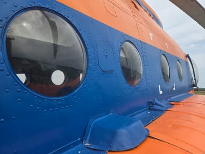 A close-up, low-angle exterior shot shows the blue and orange fuselage of a helicopter parked outdoors. The blue section features several round porthole windows, with the closest one reflecting a cloudy sky and the rotor blades. Rivets are visible along the seam where the blue and orange panels meet, and on the blue panel itself. A small, rectangular access panel with a latch is also present on the blue section. The orange section below is a flat, textured surface. A part of the helicopter's tail boom or a similar appendage extends into the upper right corner of the frame, appearing to be a light gray color. The background is a hazy, light-colored sky with some clouds. The helicopter is situated in an open outdoor area, possibly an airfield or a field, with no visible structures or immediate surrounding cityscape.