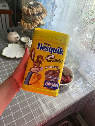 A hand with bright pink nail polish and a delicate gold chain bracelet holds a bright yellow, cylindrical plastic container of Nestlé Nesquik chocolate milk powder. The container is oriented diagonally in the frame, obscuring part of the background. The label on the container is predominantly yellow with blue and brown text and graphics. Prominently displayed is the Nestlé logo and the "Nesquik" brand name. Below this, text in Turkish reads "sütü sevdirir!" which translates to "makes milk lovable!". Further down, there is text mentioning "D Vitaminli" (with Vitamin D) and "Vitaminli Çikolatalı İçecek Tozu" (Vitamin Chocolate Drink Powder), along with nutritional information, including "420ge". A cartoon image of the Nesquik bunny character is featured on the label, appearing to pour chocolate milk from a bag.
The background shows a patterned tablecloth with a grey and white checkered design. To the left of the Nesquik container, a silver decorative object with a coiled, dough-like appearance is visible, possibly a lamp or candle holder. A small white bowl filled with red strawberries is also present in the background. Further to the left and below are other decorative ceramic items, including a white bird-like figure. The lighting suggests it is daytime, with natural light illuminating the scene. The overall impression is a casual, domestic setting, possibly a kitchen or dining area.