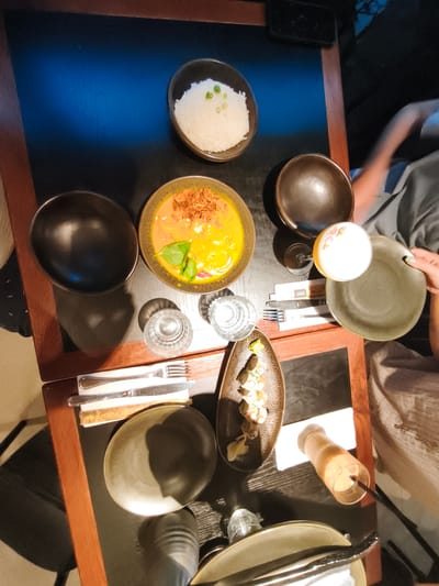 This image captures a table setting at a restaurant in Bnei Dror, Israel, showcasing a meal for at least two people. The table is a dark wood surface with a visible blue tint from lighting, set with various dishes and drinks. In the foreground, a dark, oval-shaped plate is positioned with silverware. To the left, another dark, deep bowl is empty. Above these, a lighter brown, oval dish contains a portion of rice topped with green onions. Adjacent to the rice is another deep bowl holding a vibrant orange curry-like dish garnished with fried shallots and a basil leaf.

To the right of the curry, a tall glass of a light-colored beverage with a frothy top, possibly a cocktail or creamy drink, sits next to a dark, round bowl. Further to the right, a hand with a light-colored sleeve and a green nail is reaching for a flat, greyish-green plate. Below the drinks and curry, a small, elongated dish contains pieces of sushi or similar rolled food items, accompanied by pickled ginger. Two glasses of water are also visible on the table, along with another beverage in a clear glass with an orange liquid. The background is blurred but suggests a dimly lit, indoor environment, possibly a restaurant interior, with hints of people's clothing and arms. The overall impression is of a shared dining experience with a diverse array of food and drinks.