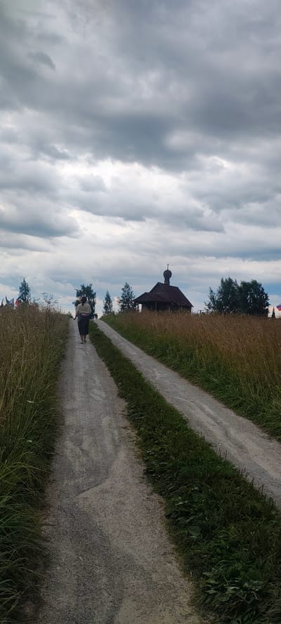 A single individual walks away from the camera down a gravel path. The person is wearing dark pants and a light-colored top, with a pink fanny pack around their waist. The path is flanked by tall, dry grass on the left and more vibrant green grass on the right. In the distance, at the end of the path, a small, dark wooden chapel with a domed roof and a cross on top is visible. The sky is overcast with heavy, gray clouds. The scene appears to be in a rural or semi-rural setting, possibly near the city of Novy Izborsk, Russia, given the presence of the chapel.