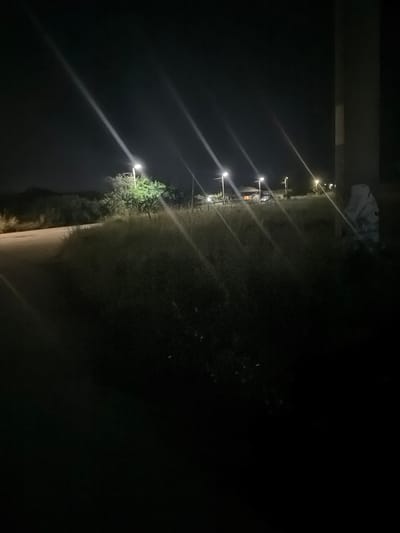 The image depicts a nighttime scene in an outdoor environment, likely a rural or suburban area. Several streetlights are illuminated, casting long streaks of light across the dark sky and illuminating the surrounding vegetation and a few buildings in the distance. The ground appears to be a mixture of dirt and grass. A portion of a large, dark, vertical structure, possibly a pillar or a utility pole, is visible on the right side of the frame. In the far distance, a vehicle is visible next to one of the buildings, and lights are on inside some of the structures. The overall impression is one of a quiet, dimly lit night. The location is identified as Smyadovo, Bulgaria.