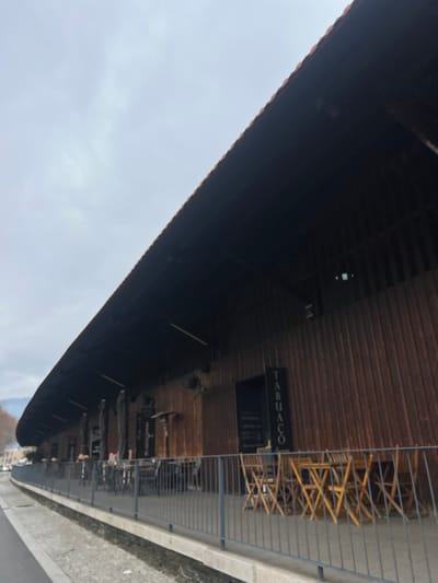 An outdoor scene in Peso da Régua, Portugal, depicts a long, single-story establishment with a steeply sloped, dark tiled roof. The building facade is constructed of vertical reddish-brown wooden planks. A prominent black vertical sign on the facade displays white text, partially legible as "TABUACO."

In front of the building, an outdoor dining area is situated on a raised concrete platform, secured by a dark grey metal railing. This area contains multiple dining arrangements: several tables with dark chairs, some positioned beneath retracted dark parasols, and a cluster of eight light-colored wooden folding chairs arranged around unseen tables in the foreground. Two dark, cylindrical patio heaters are visible along the wooden facade.

The ground immediately adjacent to the railing on the left is a paved surface, possibly a sidewalk or narrow street. The sky is uniformly overcast and light grey. In the distant background to the left, blurred hills or mountains with minimal vegetation are discernible under the horizon. No individuals are present in the frame, and no actions or interactions are occurring.
