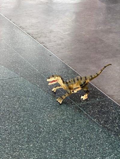 A toy velociraptor is positioned on a polished floor made of two different colored sections of terrazzo tile, divided by a thin metal strip. The left section of the floor is a dark grey with white flecks, while the right section is a lighter grey with subtle variations in tone. The dinosaur is facing left, with its mouth open and its tail curved upwards and to the right. The lighting suggests an indoor environment, possibly a public space or walkway, with a shadow cast by the dinosaur. The image is a high-angle shot, looking down at the scene. There is no information in the image to determine its location in Leeds, United Kingdom.