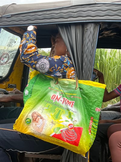 A woman in a patterned long-sleeved shirt and a gold watch is leaning against the roof of an open-air vehicle. She is holding a large green and yellow bag with the text "ON-TIME PLANT FIBER MOSQUITO COIL" printed on it. The bag displays an image of a woman holding a baby and illustrations of mosquitos and a mosquito coil. The vehicle appears to be a tuk-tuk, and the scene is set against a backdrop of tall green grass, suggesting a rural or semi-rural location, potentially in Orifite, Nigeria. Another person's arm is visible in the background. The front windshield of the tuk-tuk is cracked.