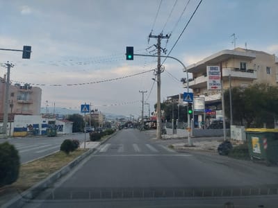 This image displays an urban street scene in the Municipal Unit of Acharnes, Greece, under an overcast sky with hints of blue. The perspective is from a low vantage point, looking directly down an asphalt road. The road features clearly marked white pedestrian crossing stripes in the immediate foreground and a narrow, central median strip with sparse low-lying vegetation.

On the right side of the road, a prominent utility pole supports numerous overhead wires, a traffic light displaying a green signal for vehicular traffic, and a smaller pedestrian traffic light also illuminated green. Adjacent to this pole, a light-colored, multi-story building with balconies exhibits a large sign in Greek for "ΞΕΝΕΣ ΓΛΩΣΣΕΣ ΚΟΝΔΕΑΚΗ" (Foreign Languages Kondeaki), complete with the phone number 210 2441416 and a red directional arrow pointing right. A large green municipal waste bin and a black plastic refuse bag are present on the far right sidewalk.

On the left side of the road, a multi-story residential building with multiple balconies is visible behind a lower structure featuring a distinctive blue and yellow painted exterior, possibly a business, with a smaller green waste bin positioned near its front. Further down the street, additional buildings, utility poles, and several parked vehicles are discernible along the road. The background reveals distant hills or mountains beneath the pale sky. No individuals are visible in the scene.