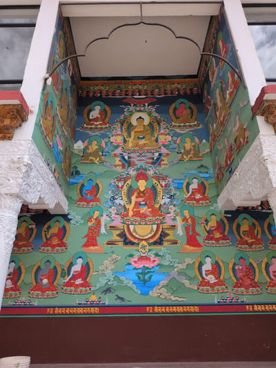 The image shows a brightly colored mural painted on the exterior wall of a building, likely a monastery or temple, in Phyang, India. The mural depicts a series of Buddha figures in various poses, seated on lotus thrones and surrounded by colorful halos. The background is a vibrant green, with stylized clouds and water features. The artwork is detailed and intricate, with each figure individually rendered with distinct colors and features. The overall impression is one of spiritual devotion and artistic mastery, common in Buddhist art.