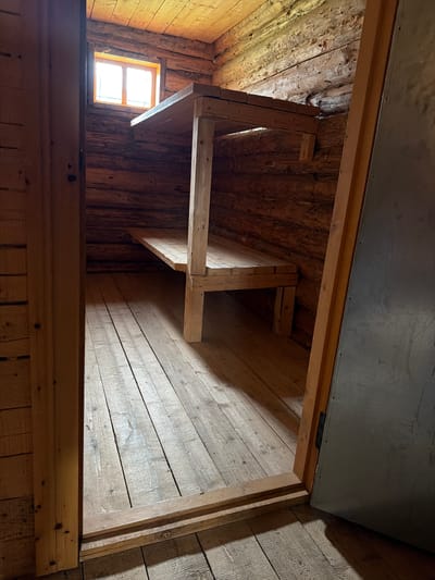 The image depicts the interior of a rustic wooden structure, likely a cabin or shelter, in Ust'-Pyzas, Russia. The walls and ceiling are constructed from rough-hewn logs, and the floor is made of wooden planks. A simple, multi-tiered wooden bunk bed system is the dominant feature within the space. Sunlight streams through a small, two-pane window positioned high on the left wall, illuminating the dust motes in the air and casting shadows. The entrance to the structure is visible on the right, with a dark, metallic door ajar, revealing a portion of the interior. The overall impression is one of a sparsely furnished, utilitarian dwelling, possibly used for basic accommodation in a remote or natural setting. There are no people or animals visible in the scene.