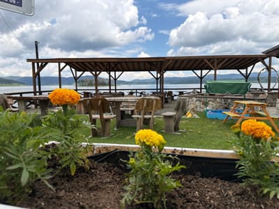 This detailed image captures an outdoor leisure area in Цигов чарк, Bulgaria, under a partly cloudy sky. In the immediate foreground, a white planter box contains dark soil with three marigold plants; two orange-flowered plants are visible on the left, with one in sharp focus, and a yellow-flowered plant is on the right. The midground reveals an open-air dining or relaxation space featuring numerous rustic wooden tables and benches/chairs, some incorporating stone bases, arranged on a manicured lawn. A substantial dark brown wooden pergola with a slatted roof provides shade over a portion of the seating area. To the right, a low stone wall supports a green fabric-covered object, likely outdoor equipment. Adjacent to this, a large white heart-shaped decorative element, possibly floral, stands near the water's edge. Further right, a small blue plastic sandbox and a multicolored wooden children's picnic table are present. The background features a large, calm body of water, consistent with the Batak Reservoir, extending to a horizon line defined by distant forested mountains. The sky above is a mix of bright blue patches and large, white cumulus clouds. No people are visible in the scene. A partial white sign with the text "09 =" is present in the upper left corner.