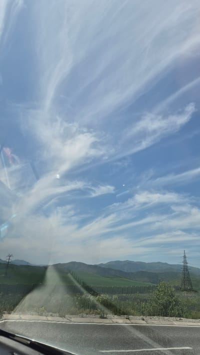 The image shows a view from a vehicle, looking out over a landscape. The sky is blue with wispy white clouds. In the distance, there are rolling green hills and mountains. A power line tower is visible on the right side of the frame. The foreground shows a road with a white line, and a metal fence running along the side of the road. The scene appears to be in a rural area. The provided location of Berezovka, Kazakhstan, suggests this is likely a landscape shot from a highway or road in that region. There are no people visible in the image.