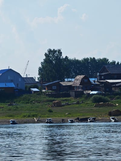 The image shows a village on the bank of a wide river. The village consists of several wooden houses, some with blue roofs. In the background, there is a dense forest. Several small boats are moored on the riverbank. The sky is partly cloudy. The scene appears to be in a rural area, possibly in the region of Verkhne-Pashino, Russia, given the architecture and natural landscape. There are no people visible in the image.