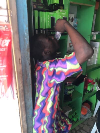 An individual is depicted standing inside a commercial establishment in Ijoko, Nigeria. The person, with dark skin and short dark hair, is wearing a long-sleeved garment featuring a distinct pattern of purple, blue, red, and lime green wavy stripes. They are observed looking upwards, actively drinking from a transparent plastic sachet held to their mouth with their right hand. Their left hand holds a dark-colored smartphone, positioned at waist level. The immediate background consists of green shelving units, densely stocked with numerous uniformly packaged items, predominantly green boxes, some displaying the "oraimo" brand. Other merchandise, including white and red packaged goods, is also visible. To the left, a blue-painted wooden doorframe or partition partially frames the scene, adjacent to which is a red sign with partially visible text, including "fer" and "ics," suggesting a transfer service or electronics. The floor is tiled with small, dark, possibly mosaic-style tiles.