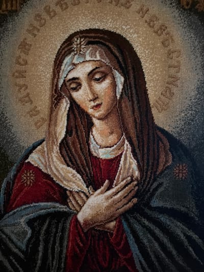 The image depicts a rug or tapestry featuring a portrait of the Virgin Mary. Her face is pale, with closed eyes and a serene expression. She is depicted wearing a dark brown head covering with a lighter-colored trim and a small, flower-like decoration. She is clad in a red garment, draped with a beige cloth and a dark blue cloak. Her hands are clasped together in prayer. The portrait is surrounded by a circular halo of lighter shades, containing text in a Slavic language. The background is a mix of dark and light shades, suggesting a neutral setting. There is no information to suggest this is in the city of Levkovo, Russia.