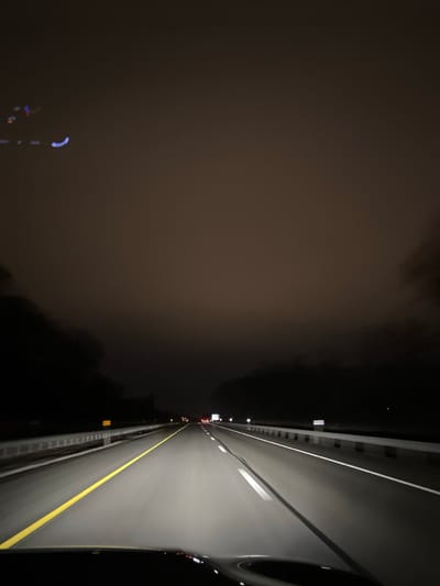 A nighttime, sideways-oriented view from a car on a highway. The car occupies the right side of the image, illuminated by headlights. A yellow line marks the road's edge. To the left is a dark, foggy expanse, obscuring visibility. Streetlights emit a faint, blurred glow through the fog. The road appears to stretch into the distance, with other vehicles visible far ahead. The overall mood is ominous, with the fog creating a sense of isolation.