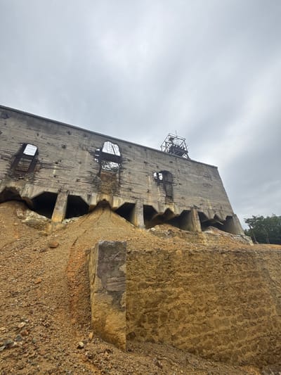 The photograph captures a low-angle view of a derelict concrete industrial structure, likely an old mine building, set against a cloudy sky. The building is weathered and damaged, with broken windows, exposed rebar, and significant erosion. Piles of earth and gravel surround the base of the structure, obscuring some of its features. The landscape appears arid and industrial. This structure is located in the city of Kapan, Armenia, which is known for its mining industry. There are no people visible in the image.