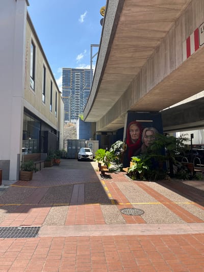 An urban scene depicts an alleyway or underpass in South Brisbane, Australia, bathed in bright daylight under a clear blue sky with sparse clouds. The image is taken from a slightly elevated perspective, looking down the length of the alley.

On the left, a massive concrete overpass structure dominates the upper part of the frame, showing weathered surfaces and a clear height limit sign displaying "2.7" in red and white. Beneath the overpass, on a shadowed wall, is a large mural depicting two faces: an older woman in a red hood or garment, and a younger individual with lighter hair, both looking forward with neutral expressions. Numerous potted plants and lush greenery are arranged along the base of this wall, adding a touch of nature to the concrete environment. Further down the alley, a multi-story building with a grid-like window pattern is visible in the distance against the sky. A white car is parked further into the alleyway.

The right side of the image features a pedestrian pathway constructed from alternating sections of reddish-brown brick pavers and lighter, speckled concrete. A thin yellow line runs along one of the concrete strips. A circular metal manhole cover is embedded in the pathway towards the middle, and a rectangular metal drainage grate is visible closer to the foreground. Strong sunlight casts distinct shadows on the pathway, indicating midday or early afternoon. The overall impression is one of a vibrant, utilized urban space with street art and greenery contrasting with industrial infrastructure.