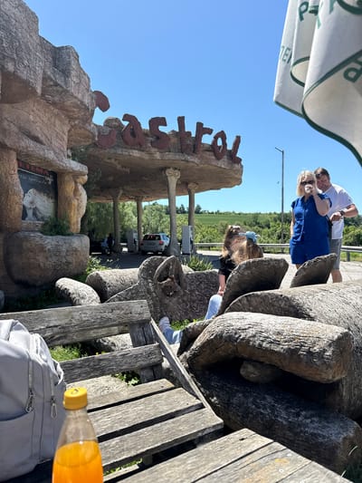 The image shows a sunny outdoor scene near a tourist attraction labeled "Sestrimo," possibly located in or near Belomortsi, Bulgaria. The name is displayed in large, rustic lettering atop a rock-like structure reminiscent of a prehistoric or cave theme.
A family of three - a man, a woman, and a young girl - are positioned near the rocks. The woman is blonde and wearing a blue dress; the man has a white shirt, and the girl has a blue hair tie. They appear to be posing or interacting with the rocks.
In the foreground, there's a partially visible wooden bench with a backpack and a bottle of orange juice. The scene is bathed in sunlight, creating bright shadows and highlighting the textures of the rocks and the objects on the bench. The background features a road with vehicles and green vegetation, suggesting a roadside location.