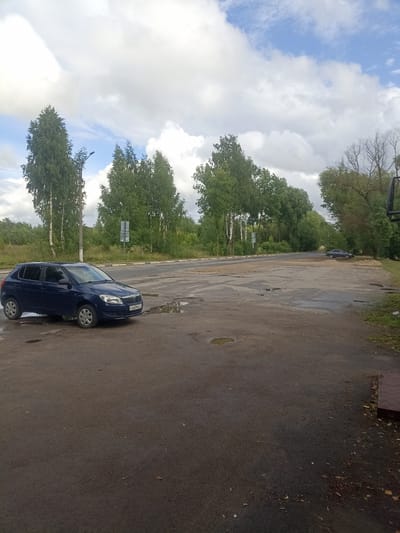 The image depicts a scene on a cloudy day at an outdoor location in Furmanov, Russia. In the foreground, a dark blue Skoda Fabia hatchback is parked on a wet asphalt surface. The car has a license plate that appears to read "XE660ME". The asphalt surface is uneven and shows signs of wear, with several puddles of water scattered across it.
In the middle ground, a road is visible, and further back, a line of trees, predominantly birches and other deciduous trees, forms a dense treeline. There are also some utility poles and traffic signs visible along the road. In the distance, a darker colored car is parked off to the right of the road.
The sky is filled with a mixture of white and grey clouds, indicating overcast or partially cloudy weather. The overall impression is of a somewhat rural or suburban environment, possibly a parking area or the side of a road, with evidence of recent rain.