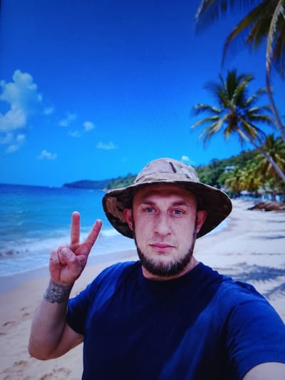 A man wearing a camouflage bucket hat and a dark blue t-shirt is posing for a selfie on a beach. He has a beard and is making a peace sign with his left hand. The background shows a sandy beach, palm trees, the ocean, and a blue sky with some clouds. The man has a tattoo on his left forearm. There is no information available about the city of Safonovo, Russia.