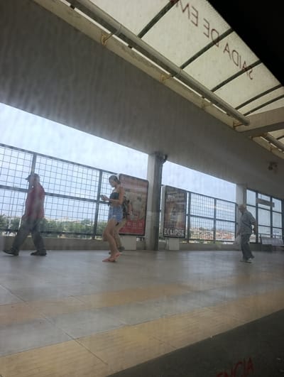 This image captures a scene at a train or bus station in Seixal, Portugal. The primary subjects are three individuals walking along a tiled platform. On the left, a man wearing a red shirt, dark pants, and a cap walks away from the camera. In the middle, a woman with dark hair, wearing a black top and denim shorts, is looking at her phone. She has a backpack on. On the right, an older man in a gray shirt and pants walks towards the right of the frame, carrying a white plastic bag.
The setting is an outdoor or semi-outdoor covered area, indicated by the translucent roof with visible beams and pipes overhead, and a metal grid fence bordering the platform. The time of day appears to be daytime, with bright, possibly overcast, sky. The overall atmosphere seems ordinary, with people going about their daily commute.
Several pieces of visible text provide context. On the roof, red lettering is partially visible, appearing to say "SAÍDA DE EV" which likely translates to "EXIT FROM EV" or similar. On posters along the platform, text in Portuguese is visible, including "OS TEUS SABORES DE VERÃO COM 5 ZEROS NO DECE", "Yoggi", "Quando os astros sedilham", "ECLIPSE", and "CORRIOS" (Correios, meaning Post Office). The presence of "CORRIOS" suggests the station may be near a post office or part of a larger transportation hub. The tile pattern on the platform is a mix of beige and grey squares, with textured strips in some areas, possibly for tactile guidance.