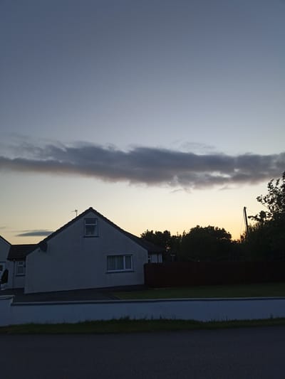 The image depicts a residential scene in Harley Court, Ireland, during twilight. A two-story house with a white facade and a pitched roof is visible, featuring windows with blinds. Adjacent to it, another house with a tiled roof is partially seen. A dark brown wooden fence runs horizontally across the middle ground. Silhouetted trees are present in the background, and a power pole stands to the right. The sky is a gradient of muted blues and yellows, with a horizontal band of dark clouds. The foreground shows a paved road and a white rendered wall with grass. No people are present in the image.