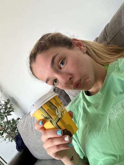 The image captures a young woman indoors, likely in Harish, Israel, looking directly at the camera with a playful, slightly pouting expression. Her head is tilted, presenting the image in a sideways orientation. She has blonde or light brown hair and blue-grey eyes.
She is wearing a light green t-shirt with white text across the chest, partially visible as "DICK," likely from a brand like "DICKIES." On her left wrist, she has a small red heart tattoo and a blue beaded bracelet. Her left ear features two small silver hoop piercings in the upper cartilage.
In her right hand, she holds a yellow coffee cup adorned with a green cactus design. Visible text on the cup includes "CAFE." Her fingernails are painted, with one nail being white and the others a light turquoise blue.
The woman appears to be seated on a gray upholstered couch or chair, which is visible behind her. A potted plant with green leaves, possibly eucalyptus, is visible in the lower right foreground. The background consists of a bright, plain white wall or open space, indicating a well-lit indoor environment. The overall mood conveyed is casual and relaxed.