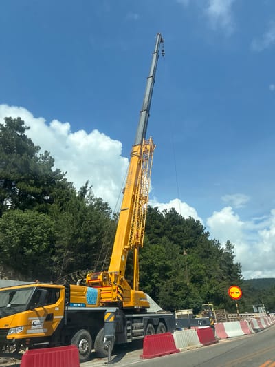 A yellow mobile crane is parked on the side of a road in Sosnovy, Russia, near a wooded area. The crane's boom is extended high into the air. Construction barriers, including red and white plastic barriers, line the road. A traffic sign depicting two cars with a red circle and diagonal line over them, indicating no overtaking, is visible. The sky is blue with white clouds. The crane has branding that appears to be in Russian, including "СТРОЙ ТРАНС" and "XCT10.5".