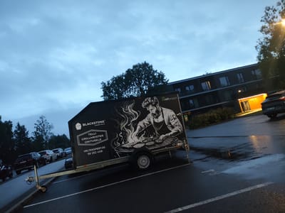 The image depicts a mobile grill trailer parked in a parking lot, likely in Tiskre, Estonia, based on the language on the trailer. The trailer has a large graphic of a man grilling on one side, with text that reads "BLACKSTONE CERAMIC GRILLS" and "TELLI GRILLMEISTER ÜRITUSELE VELTEKSPERT.EE 56 252 408". The background shows a modern building with several windows, trees, and a cloudy sky. Several cars are parked in the lot, and the ground appears wet, suggesting it has recently rained. The lighting indicates it is either early morning or late evening. There are no people clearly visible in the scene.