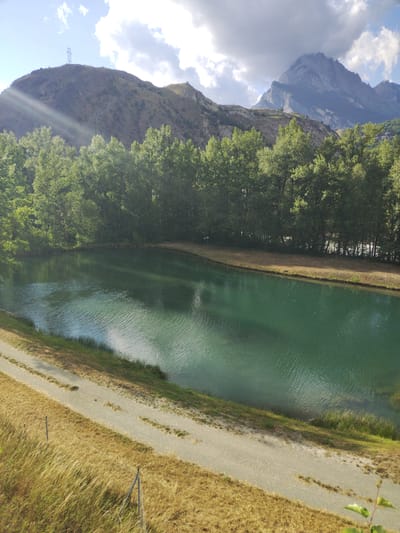 This is a wide, high-angle, full shot of a scenic landscape in Saint-Michel-de-Maurienne, France. The image captures a tranquil body of water, likely a lake or reservoir, with its surface rippling and reflecting the surrounding environment. The water is a deep, emerald green, creating a striking contrast with the dry, golden grasses that line the foreground. A gravel path winds its way along the edge of the water, suggesting a recreational area or hiking trail.
In the background, a dense forest of lush green trees forms a verdant wall behind the water. Beyond the trees, rugged, rocky mountains rise dramatically, their peaks partially obscured by dramatic, cloudy skies. Sunlight streams through the clouds, casting rays onto the mountainside and illuminating the scene with a bright, clear light. The overall atmosphere is one of peaceful serenity, a perfect summer day in a mountainous region. There is no visible text in the image.