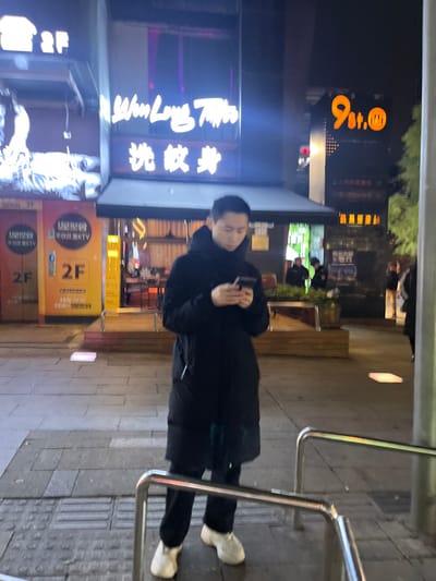 The image depicts a night-time urban street scene, centered around a person standing on a sidewalk in what appears to be Tian’gongdian Subdistrict, China. The primary subject is an adult, likely male, wearing a dark, possibly black, long winter coat or jacket and dark trousers, paired with light-colored, possibly white or cream, chunky shoes. He is holding a smartphone with both hands, looking down at it, engrossed in its use.
The setting is an illuminated street at night, with several brightly lit signs from various establishments. To the person's left, multiple neon signs are visible on a building. One prominent white neon sign displays Chinese characters that translate to "快乐限" (Happy Limit) and "洗纹身" (Tattoo removal), with "Long" also partially visible. Further back, another large orange sign shows "9st." (likely "9th street" or similar).
To the person's right, there are metal railings, possibly for crowd control or as a bicycle rack, separating the sidewalk from another area. In the background, illuminated windows and what might be a wooden bench or planter are discernible. Another building sign is yellow/orange, featuring the text "2F" (indicating the second floor) and Chinese characters "不仅仅是KTV" (More than just KTV) and "星型管" (Star-shaped tube, or relating to KTV). The overall atmosphere is that of a bustling urban area after dark, characterized by artificial light from commercial signs. The person's expression is one of focus on their phone.