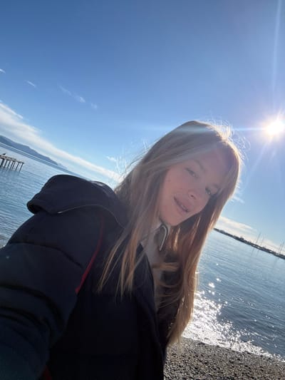 The image displays a close-up selfie of a young female subject standing on a pebbly beach in Santa Margherita Ligure, Italy. The subject, positioned centrally and angled slightly to her right, has long, light brown hair with bangs and wears a dark blue puffer jacket with a red stripe detail, layered over a light-colored turtleneck. Her lips are slightly parted, and she exhibits a faint smile.
The scene is set on a sunny day under a clear, bright blue sky with minimal white wispy clouds. The sun is visible in the upper right quadrant, casting a prominent lens flare and illuminating the water's surface to the right, creating sparkling reflections. To the subject's left, a wooden pier structure, consisting of multiple dark vertical posts, extends into calm blue-green water. A dark-colored bird is perched on one of these posts. Distant, light-colored mountains or hills are visible across the water in the background. To the subject's right, beyond the sparkling water, a grouping of boats with masts is discernible, indicating a marina or harbor area.