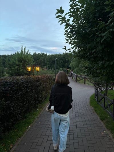 A woman with shoulder-length brown hair, wearing a black jacket and light blue jeans, walks away from the camera down a brick pathway in Izmailovo, Russia. The pathway is bordered by a dark green hedge on the left and a wooden fence on the right. Two ornate street lamps with warm, yellow light are visible behind the hedge. A dense forest of green trees forms the background, with a wooden bridge spanning across a slight incline to the right of the pathway. The sky is overcast and appears to be dusk.