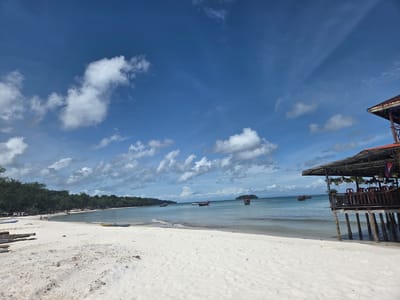 The image depicts a serene beach scene in Koh Toch Village, Cambodia, under a bright blue sky with scattered white clouds. The foreground is dominated by a pristine white sand beach, with a few scattered pieces of driftwood and some small rocks. In the mid-ground, the calm, turquoise water of the sea gently laps at the shore. Several small boats, some with people aboard, are visible on the water. A long stretch of lush green trees lines the coastline in the distance. To the right, an elevated wooden structure, likely a restaurant or bar, stands on stilts over the water, featuring a thatched roof and wooden railings. The overall impression is one of a tranquil tropical paradise.