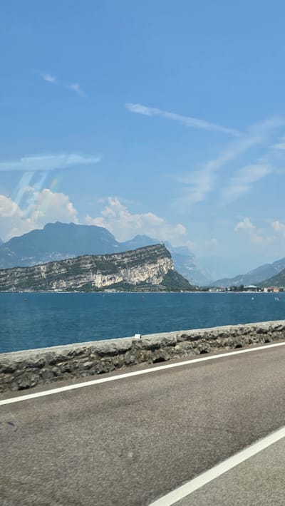 The image depicts a scenic view of a lake and mountains, likely taken from a vehicle traveling along a road. The composition is dominated by a bright blue sky with wispy white clouds and a few contrails. To the left of the frame, a rugged mountain range rises from the clear, blue water of a lake. The mountains are covered in vegetation on their lower slopes, transitioning to rocky, barren peaks higher up. Some structures appear to be built on the rocky outcrops of one of the prominent mountains.
The water of the lake is calm and reflects the blue of the sky, with small boats or vessels visible on its surface, suggesting recreational activity. On the right side of the image, a paved road with a white line marking runs parallel to the lake. The edge of the road is lined with stones or rocks, creating a natural barrier between the road and the water.
The sunlight suggests it is daytime, likely late morning or early afternoon, given the brightness and clear skies. The weather appears to be fair and sunny, conducive to outdoor activities. The overall atmosphere is one of tranquility and natural beauty. The scene is consistent with the location context of Torbole, Italy, which is known for its picturesque lake and mountain landscapes. No visible text is present in the image.
