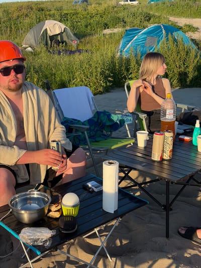 In Baltiysk, Russia, on a sandy beach with tall green grass and a clear sky, two individuals are engaged in a camping activity. In the foreground, a man wearing an orange hard hat and sunglasses is seated, wrapped in a light-colored towel. He is holding a can of what appears to be a beverage. To his right, a black foldable table displays a metal bowl, bread, a bright yellow camping lantern, a small black speaker, and a plastic-wrapped item. In the background, a woman with blonde hair is seated in a green camping chair, wearing a brown tank top and dark pants. She is looking to her right, her hands clasped. A large plastic bottle of amber-colored liquid and two beverage cans are on the table. Two tents are visible in the grassy area behind them; one is green and beige, and the other is blue. Further in the distance, two vehicles can be seen on a dirt path. The scene is illuminated by sunlight, casting shadows on the sand.
